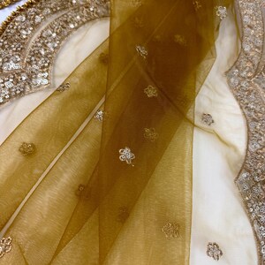 May include: Close-up of a sheer, golden-brown fabric with delicate floral embroidery. The fabric is draped over a cream-colored surface, with ornate gold sequin detailing visible along the edges. The overall aesthetic suggests a luxurious textile.