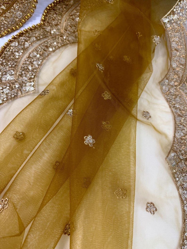 May include: Close-up of a sheer, golden-brown fabric with delicate floral embroidery. The fabric is draped over a cream-colored surface, with ornate gold sequin detailing visible along the edges. The overall aesthetic suggests a luxurious textile.