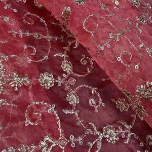 Peut inclure: Gros plan sur un tissu rouge transparent avec des broderies argent&eacute;es complexes. La broderie pr&eacute;sente des motifs floraux et tourbillonnants, avec de petits sequins ajoutant de l'&eacute;clat. Le tissu est pli&eacute;, montrant la texture et la profondeur du motif.