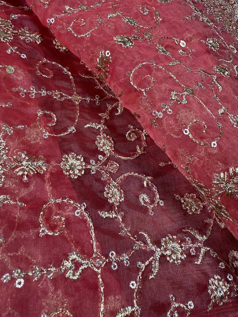 Peut inclure: Gros plan sur un tissu rouge transparent avec des broderies argent&eacute;es complexes. La broderie pr&eacute;sente des motifs floraux et tourbillonnants, avec de petits sequins ajoutant de l'&eacute;clat. Le tissu est pli&eacute;, montrant la texture et la profondeur du motif.