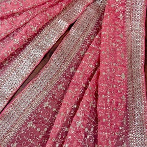 Peut inclure: Gros plan sur un tissu rose avec des broderies et des sequins d&eacute;taill&eacute;s. Le tissu transparent pr&eacute;sente de d&eacute;licats motifs floraux et une bordure scintillante. Le design est &eacute;l&eacute;gant, adapt&eacute; aux occasions sp&eacute;ciales.