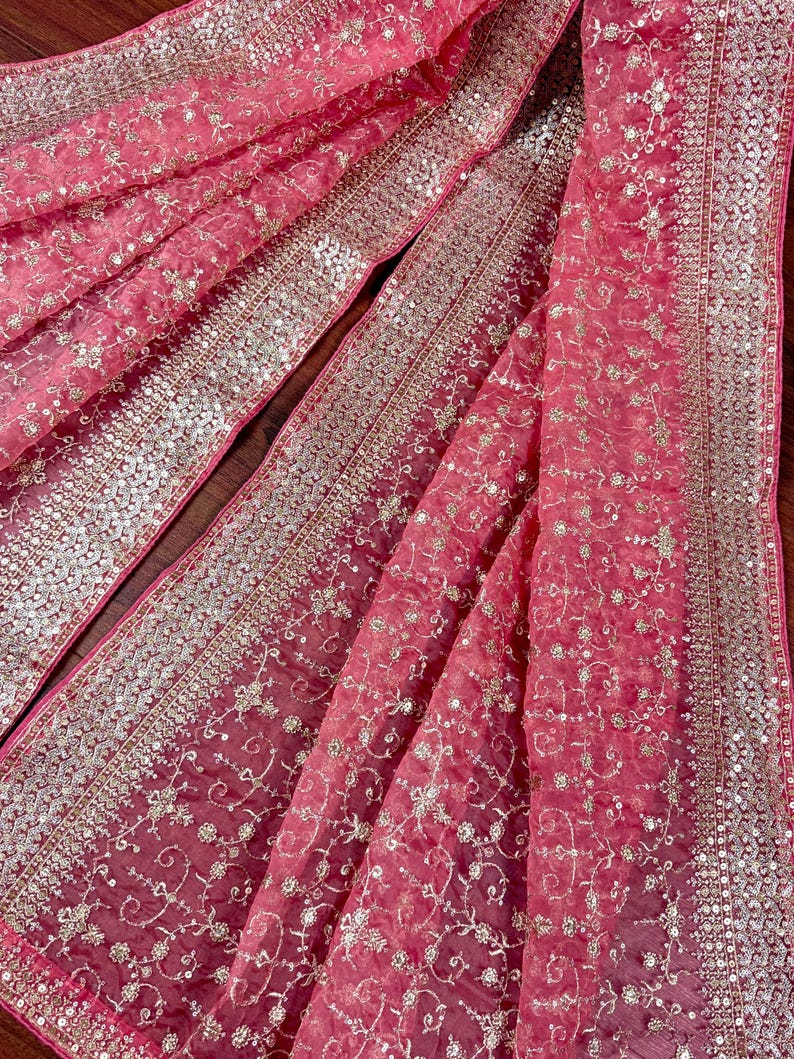 Peut inclure: Gros plan sur un tissu rose avec des broderies et des sequins d&eacute;taill&eacute;s. Le tissu transparent pr&eacute;sente de d&eacute;licats motifs floraux et une bordure scintillante. Le design est &eacute;l&eacute;gant, adapt&eacute; aux occasions sp&eacute;ciales.
