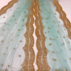 May include: A delicate, light blue fabric with gold embroidery. The edges are scalloped and adorned with gold sequins and beads. Small gold floral details are scattered across the sheer material, creating a luxurious look.