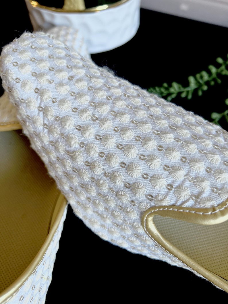 Pode incluir: Um par de chinelos brancos bordados com acabamento dourado. Os chinelos t&ecirc;m um design acolchoado texturizado.