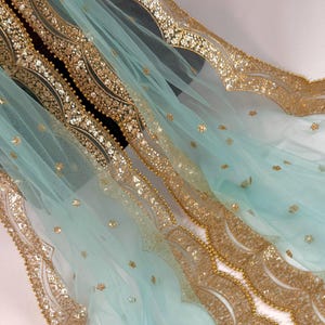 May include: A delicate, flowing fabric in a pale turquoise colour, embellished with gold sequins and beading. The edges are scalloped, creating a decorative border. The overall effect is one of luxury and elegance.