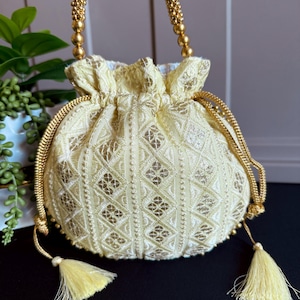 May include: A cream-coloured drawstring bag with a gold beaded handle and gold accents. The bag features an embroidered diamond pattern and is adorned with gold tassels. The bag is a small, decorative accessory.