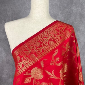May include: A vibrant red fabric with an intricate gold floral pattern. The fabric is draped over a mannequin, showcasing the detailed design and texture. The gold pattern includes floral and leaf motifs, creating a luxurious look.