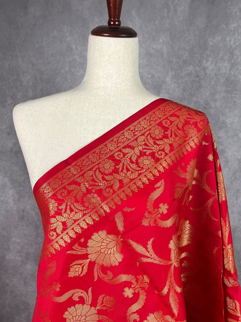 May include: A vibrant red fabric with an intricate gold floral pattern. The fabric is draped over a mannequin, showcasing the detailed design and texture. The gold pattern includes floral and leaf motifs, creating a luxurious look.