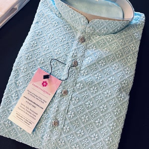 Pu&ograve; includere: Una kurta di colore azzurro chiaro ricamata con un motivo floreale bianco. La kurta ha un colletto alla coreana e una chiusura con bottone. L'etichetta sulla kurta riporta "Kaash Collection" e include le informazioni di contatto dell'azienda.