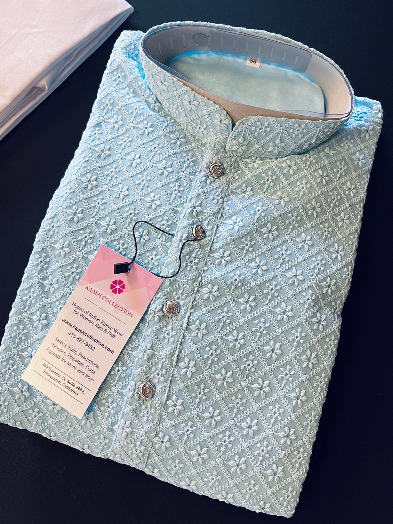 Pu&ograve; includere: Una kurta di colore azzurro chiaro ricamata con un motivo floreale bianco. La kurta ha un colletto alla coreana e una chiusura con bottone. L'etichetta sulla kurta riporta "Kaash Collection" e include le informazioni di contatto dell'azienda.
