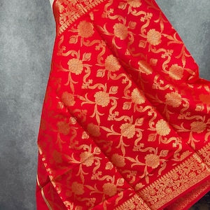 May include: A vibrant red fabric with a gold floral pattern and decorative border. The textile is draped over a form, showcasing the intricate design and rich color. The fabric's texture and details are clearly visible.