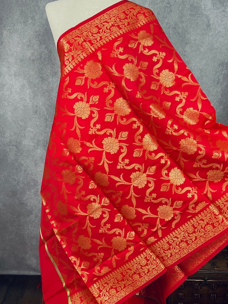 May include: A vibrant red fabric with a gold floral pattern and decorative border. The textile is draped over a form, showcasing the intricate design and rich color. The fabric's texture and details are clearly visible.