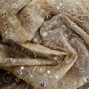 May include: Close-up of a sheer, beige fabric with intricate silver embroidery. The fabric is adorned with floral and swirling patterns, embellished with sequins. The delicate material is draped, creating soft folds and shadows, suggesting a luxurious textile.