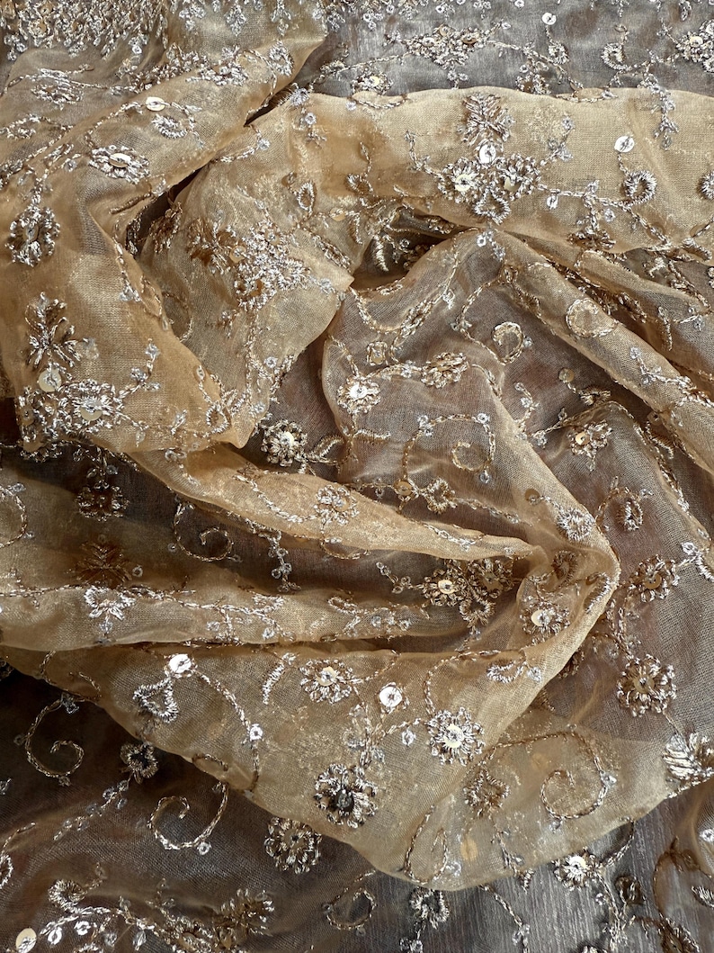 May include: Close-up of a sheer, beige fabric with intricate silver embroidery. The fabric is adorned with floral and swirling patterns, embellished with sequins. The delicate material is draped, creating soft folds and shadows, suggesting a luxurious textile.