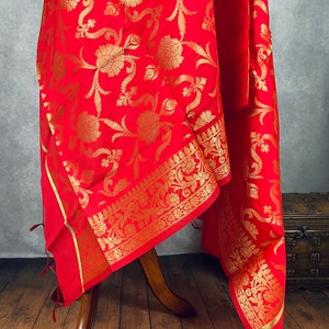 May include: A vibrant red textile with an intricate gold floral pattern. The fabric is draped over a wooden stand, showcasing the detailed design and border. The textile's rich color and ornate details suggest a luxurious quality.