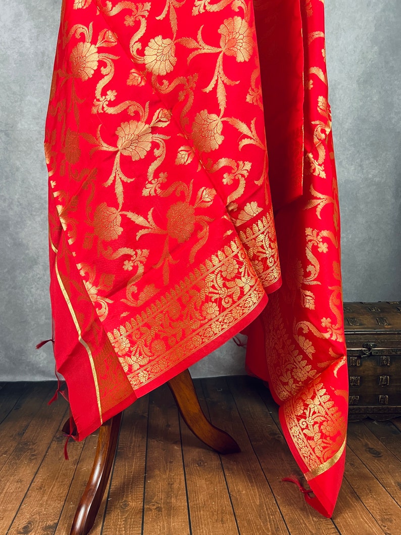 May include: A vibrant red textile with an intricate gold floral pattern. The fabric is draped over a wooden stand, showcasing the detailed design and border. The textile's rich color and ornate details suggest a luxurious quality.