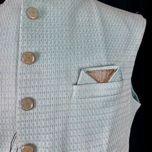 May include: A light blue sleeveless vest with a geometric pattern and brown buttons. The vest has a pocket on the left side with a brown fabric pocket square.