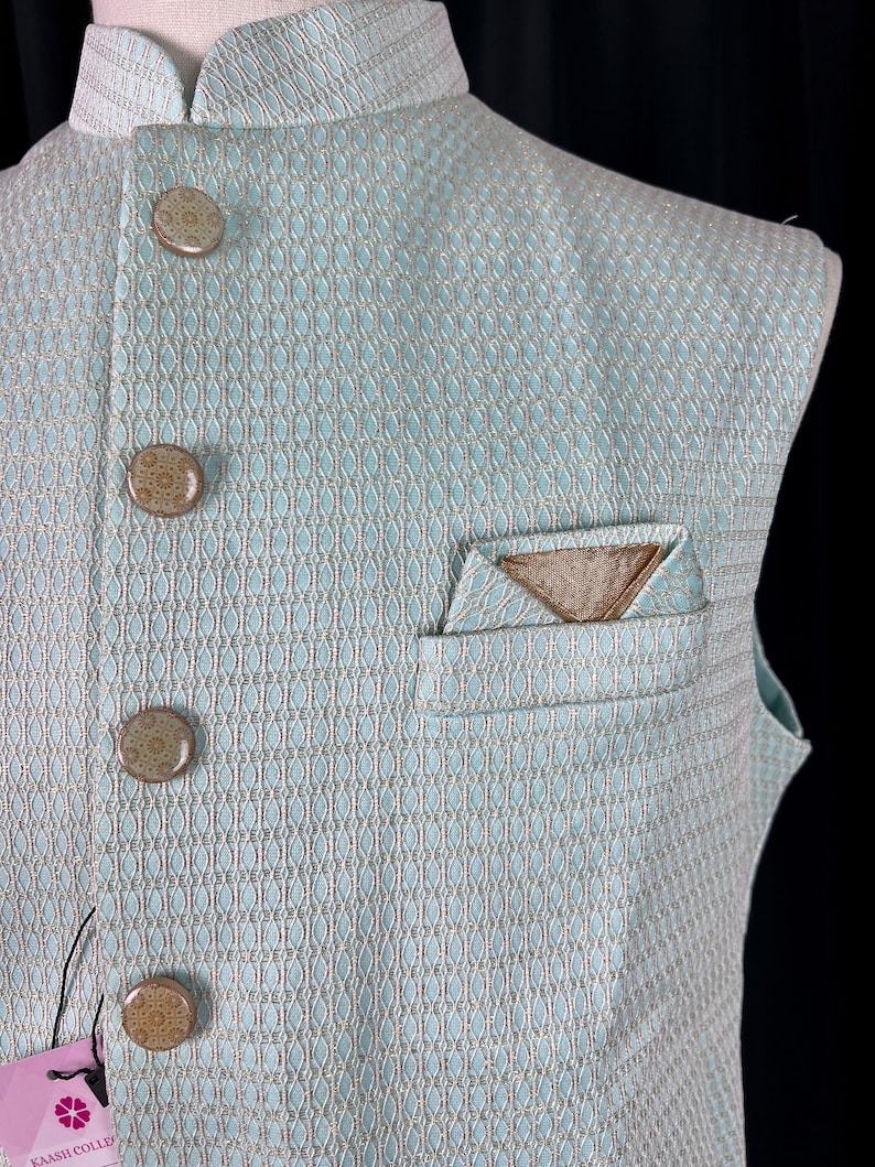 May include: A light blue sleeveless vest with a geometric pattern and brown buttons. The vest has a pocket on the left side with a brown fabric pocket square.