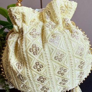 May include: A pale yellow drawstring pouch bag featuring intricate white and metallic gold geometric embroidery. The bag is accented with a handle made of large gold-toned spherical beads and includes matching pale yellow tassels at the base.