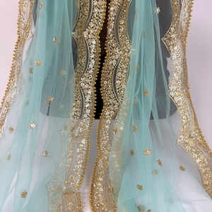 May include: A delicate, light blue fabric adorned with elaborate gold embroidery and embellishments. The borders and central design showcase detailed patterns of sequins and beads. Scattered across the surface are delicate floral accents, creating a luxurious and ornate appearance.