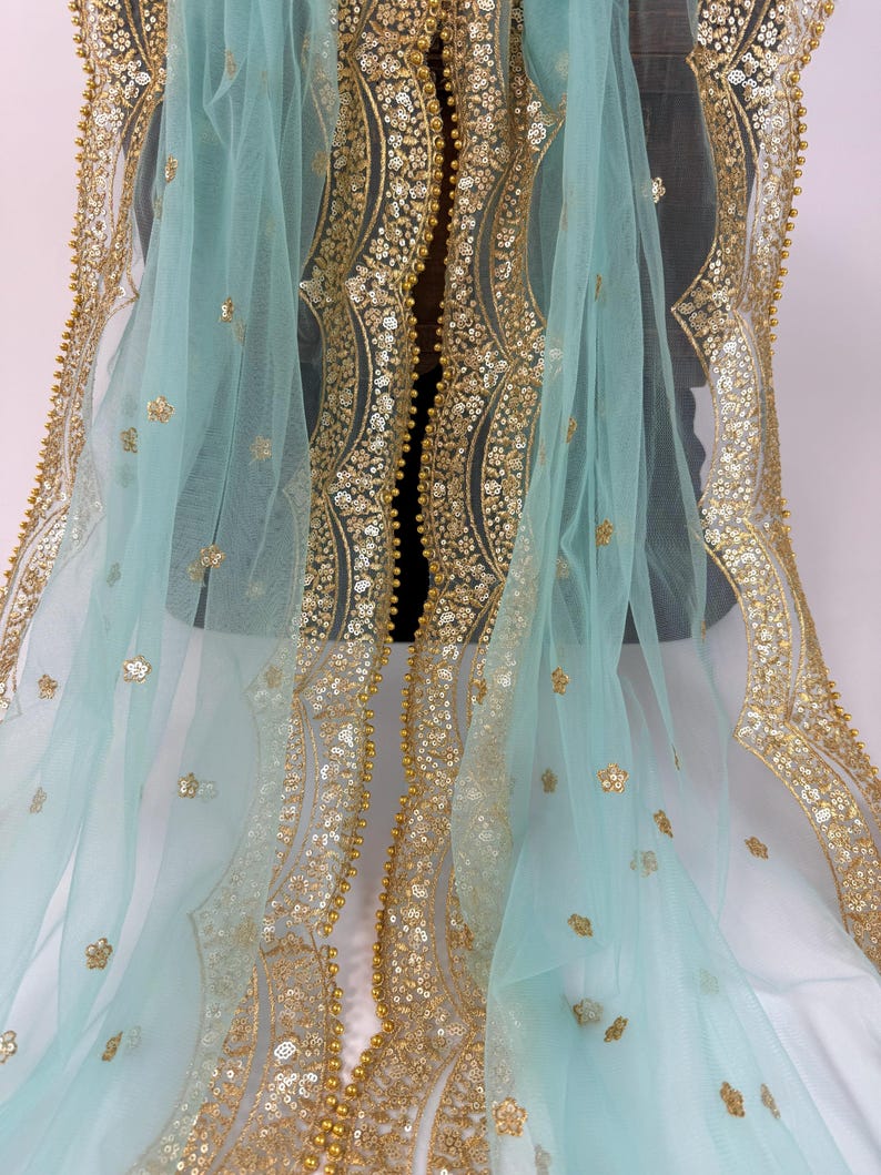 May include: A delicate, light blue fabric adorned with elaborate gold embroidery and embellishments. The borders and central design showcase detailed patterns of sequins and beads. Scattered across the surface are delicate floral accents, creating a luxurious and ornate appearance.