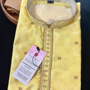 Op de afbeelding: Een gele kurta met gouden borduurwerk rond de halslijn en mouwen. De kurta heeft een klein, roze label met de tekst "KAASH COLLECTION" en contactgegevens.