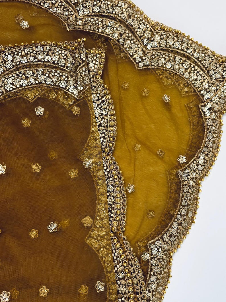 May include: Close-up of a mustard-colored sheer fabric with intricate gold embellishments. The edges are adorned with sequins and small, decorative floral patterns. The fabric has a delicate, flowing appearance.