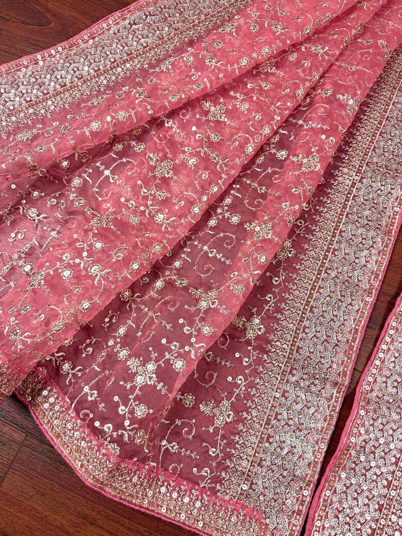 Peut inclure: Un tissu rose transparent drap&eacute; avec une broderie argent&eacute;e complexe. Le tissu pr&eacute;sente un motif floral et est bord&eacute; d'une garniture &agrave; paillettes d&eacute;taill&eacute;e. Le mat&eacute;riau d&eacute;licat est pli&eacute;, mettant en valeur sa texture et son design.