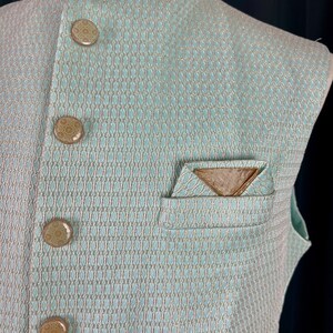 May include: A light blue sleeveless vest with a white and gold patterned design. The vest has a mandarin collar and four brown buttons. There is a pocket on the left side of the vest with a gold triangle design.