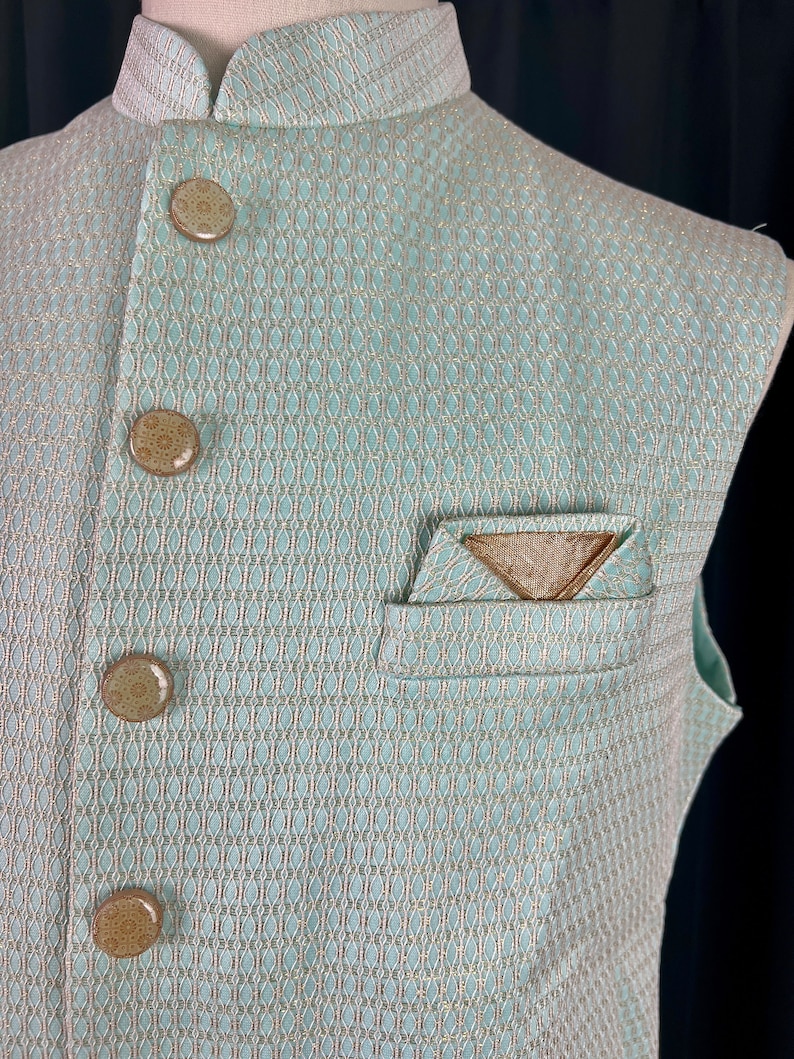 May include: A light blue sleeveless vest with a white and gold patterned design. The vest has a mandarin collar and four brown buttons. There is a pocket on the left side of the vest with a gold triangle design.