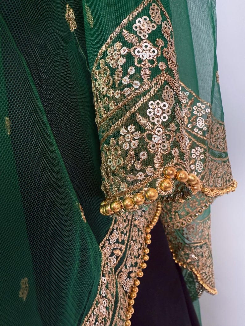 Peut inclure: Gros plan sur un v&ecirc;tement vert avec des broderies et des sequins dor&eacute;s. Les manches sont orn&eacute;es de motifs floraux et d'une bordure perl&eacute;e. Le tissu est une maille transparente, superpos&eacute;e &agrave; un tissu plus fonc&eacute;.