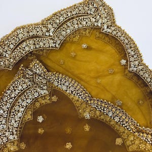 May include: Mustard-colored sheer fabric with ornate gold and silver embellishments. The edges are intricately decorated with sequins and small gold beads. Delicate floral patterns are scattered across the surface, creating a luxurious and elegant appearance.
