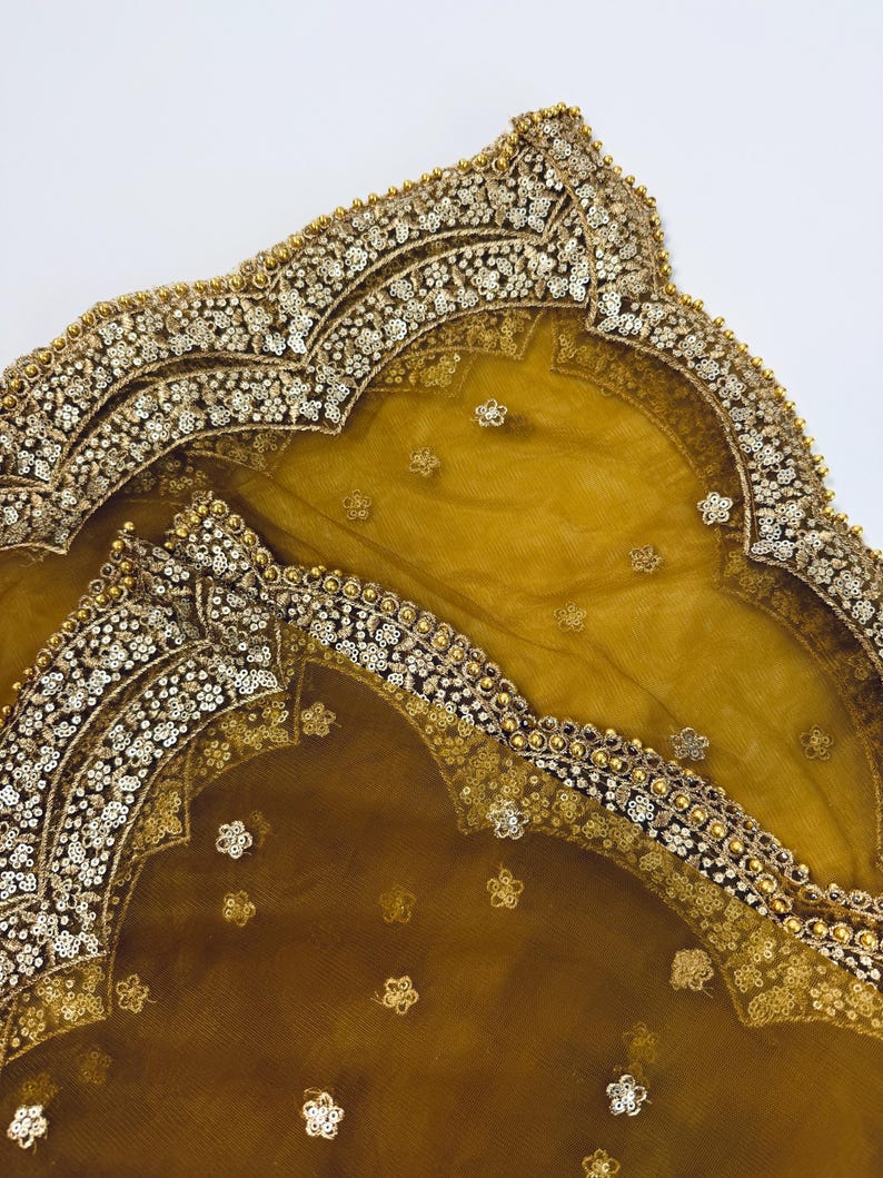 May include: Mustard-colored sheer fabric with ornate gold and silver embellishments. The edges are intricately decorated with sequins and small gold beads. Delicate floral patterns are scattered across the surface, creating a luxurious and elegant appearance.