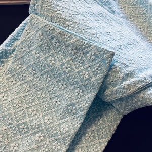Pu&ograve; includere: Un tessuto azzurro chiaro con un motivo ricamato bianco di piccoli fiori e rombi. Il tessuto &egrave; piegato a met&agrave;, mostrando il motivo su entrambi i lati.