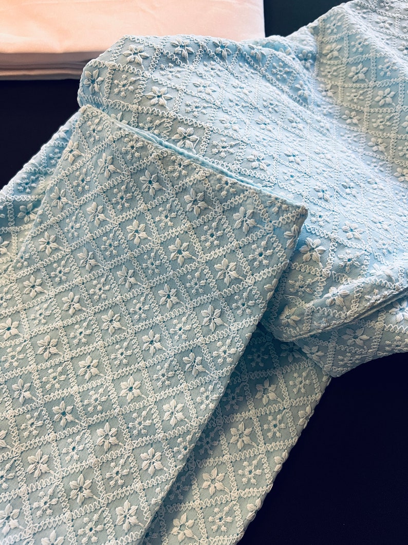 Pu&ograve; includere: Un tessuto azzurro chiaro con un motivo ricamato bianco di piccoli fiori e rombi. Il tessuto &egrave; piegato a met&agrave;, mostrando il motivo su entrambi i lati.