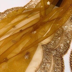 May include: Close-up of a sheer, golden-yellow fabric with delicate floral embroidery and a beaded border. The fabric is draped, showcasing its texture and the intricate details of the embellishments. The design is elegant and ornate.