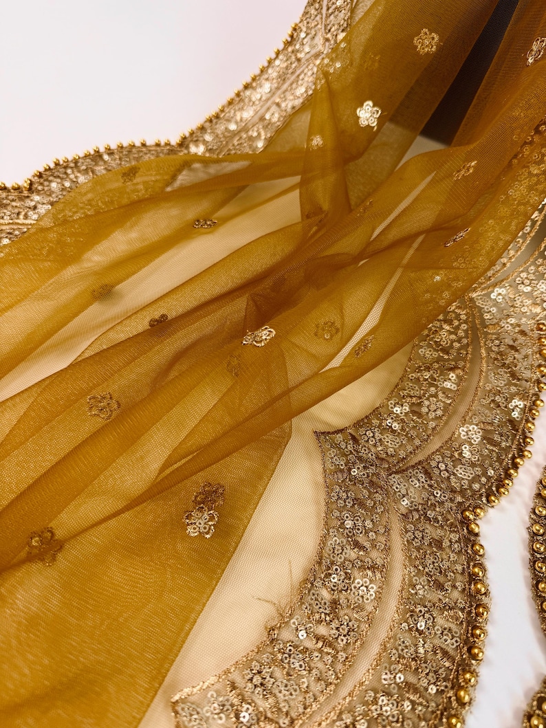 May include: Close-up of a sheer, golden-yellow fabric with delicate floral embroidery and a beaded border. The fabric is draped, showcasing its texture and the intricate details of the embellishments. The design is elegant and ornate.