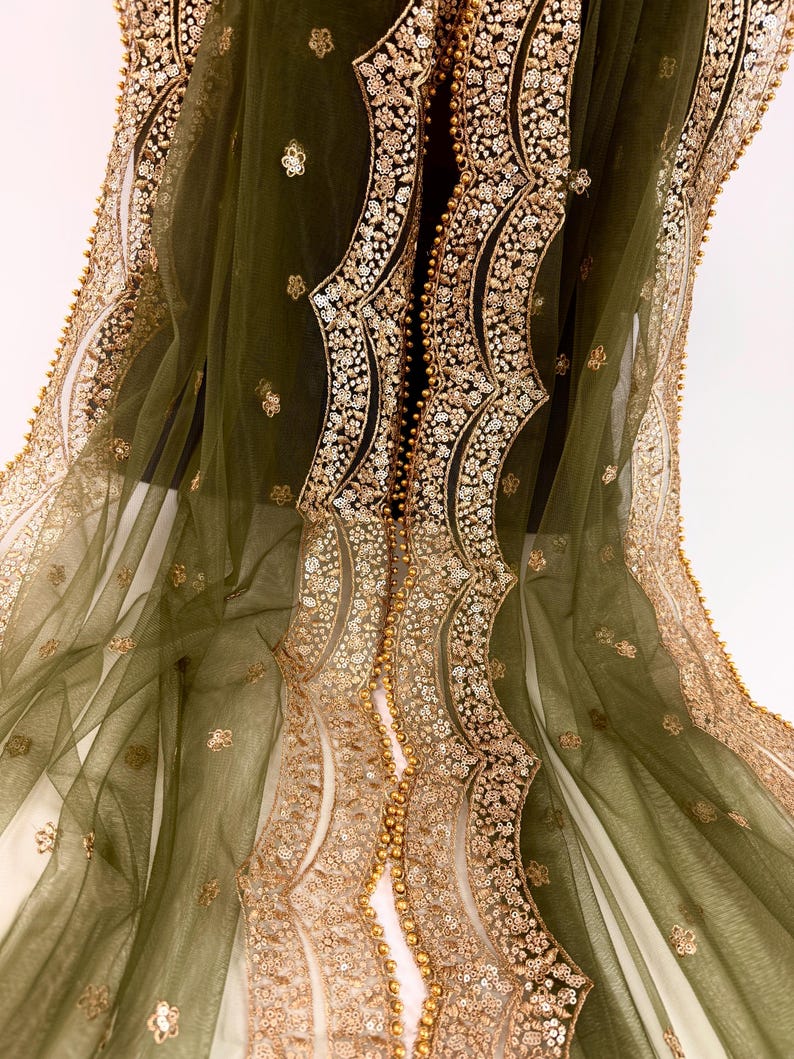May include: Olive green sheer fabric with intricate gold sequin and bead embellishments. The design features ornate, flowing patterns along the edges and center, creating a luxurious and elegant aesthetic. The fabric is draped, showcasing its delicate texture.