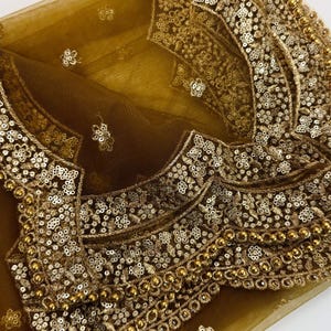 May include: Close-up of a gold-colored, intricately embroidered garment. The fabric is a sheer, golden mesh, overlaid with elaborate patterns of sequins and beads. The design features floral and geometric motifs, creating a luxurious and ornate appearance.