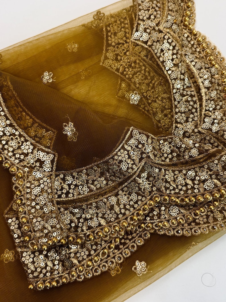 May include: Close-up of a gold-colored, intricately embroidered garment. The fabric is a sheer, golden mesh, overlaid with elaborate patterns of sequins and beads. The design features floral and geometric motifs, creating a luxurious and ornate appearance.
