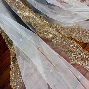 May include: White sheer fabric with a gold embroidered border. The border features a repeating pattern of floral and geometric designs.