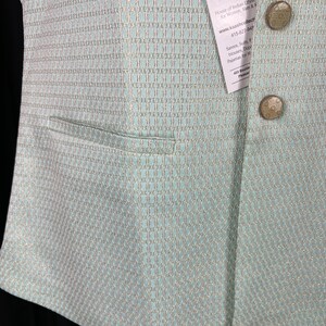 May include: A light blue vest with a gold and white patterned design. The vest has a pocket on the left side and two buttons on the right side. The vest is made of a soft, comfortable fabric. The tag on the vest reads "KAASH COLLECTION House of Indian Ethnic for Women, Men & Kids www.kaashcollect.com 415-827-948 Sarees, Suits, Blouses, Dupattas, Pajamas for Men & Women 405 Boulder Pleasanton"