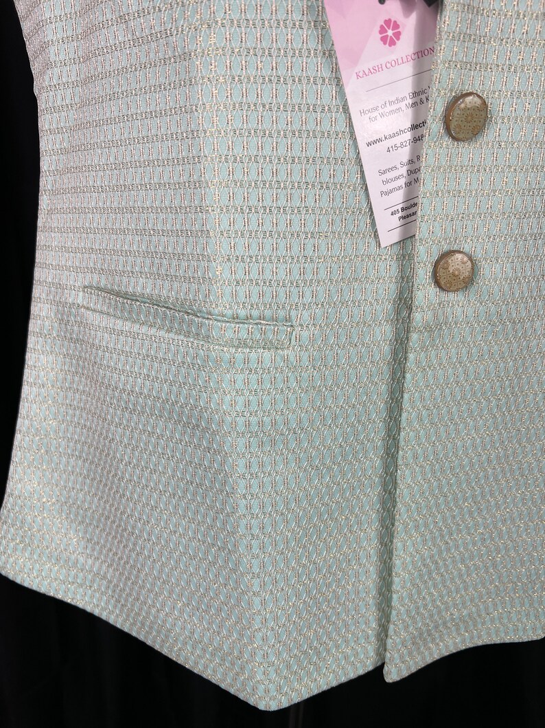 May include: A light blue vest with a gold and white patterned design. The vest has a pocket on the left side and two buttons on the right side. The vest is made of a soft, comfortable fabric. The tag on the vest reads "KAASH COLLECTION House of Indian Ethnic for Women, Men & Kids www.kaashcollect.com 415-827-948 Sarees, Suits, Blouses, Dupattas, Pajamas for Men & Women 405 Boulder Pleasanton"