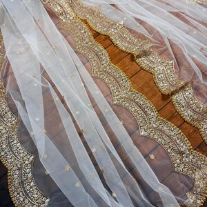 May include: White sheer fabric with a gold beaded and sequined border. The fabric is draped over a wooden surface.