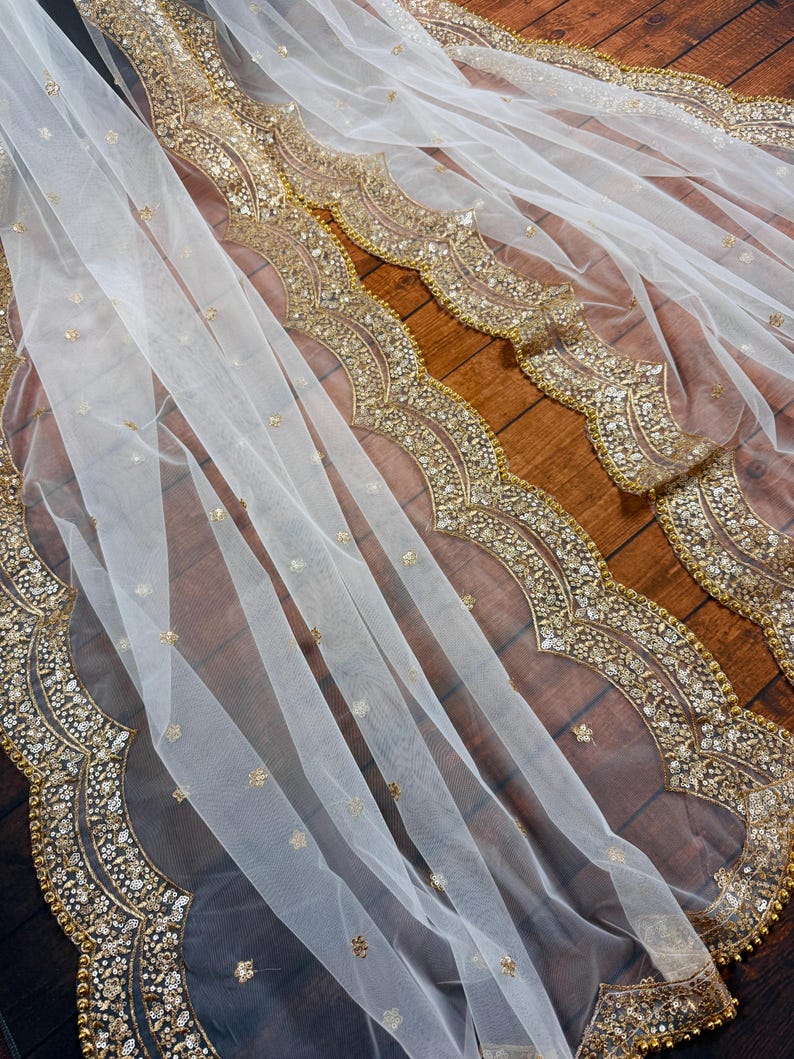 May include: White sheer fabric with a gold beaded and sequined border. The fabric is draped over a wooden surface.