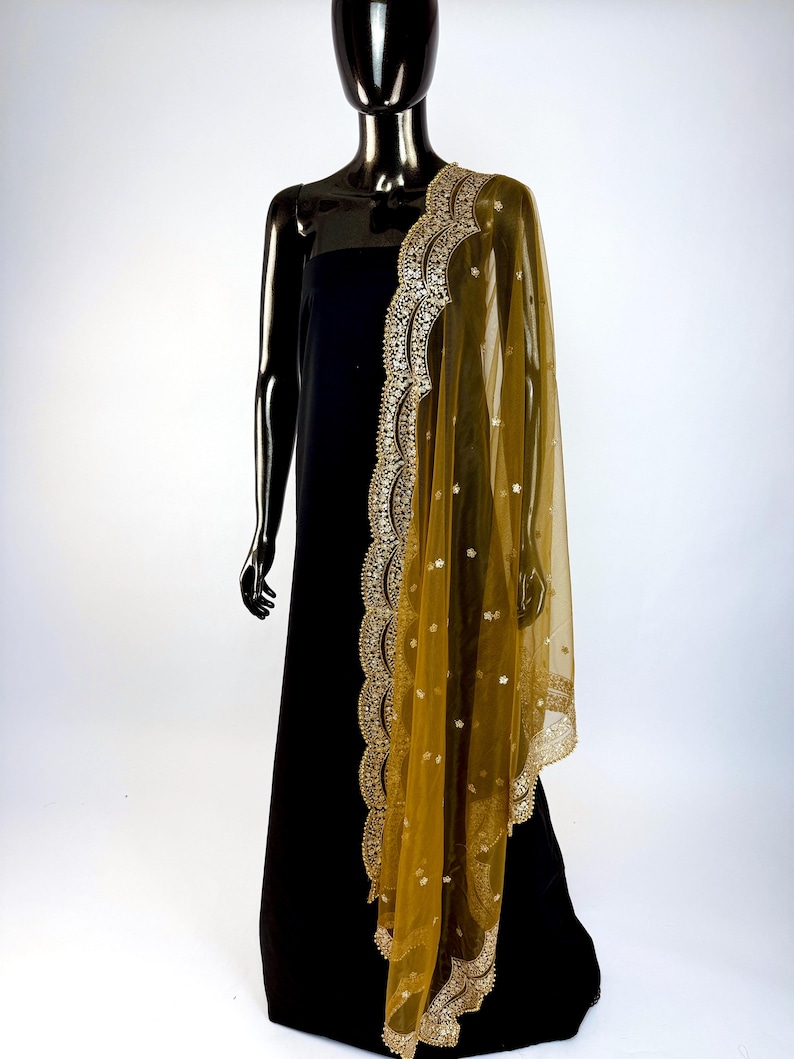 May include: A black dress is draped with a sheer, golden-yellow dupatta. The dupatta features intricate gold embroidery along the edges and scattered embellishments. The mannequin is black and stands against a white backdrop.