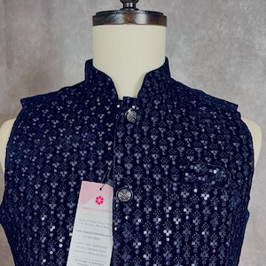 May include: A black velvet waistcoat with a mandarin collar and silver buttons. The waistcoat is decorated with a repeating pattern of sequins. The tag attached to the waistcoat reads "KAASH COLLECTION House of Indian Ethnic Wear for Women, Men & Kids www.kaashcollection.com 415-827-6482  Made in India, Handmade Suits, Dupatta, Kurta, Salwar for Men and Boys 951 Boulder Ct, Suite 204,  Pleasanton, California 94566"