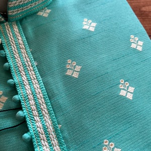 Pode incluir: Uma kurta azul turquesa com uma guarni&ccedil;&atilde;o branca e dourada. A guarni&ccedil;&atilde;o tem uma s&eacute;rie de pequenos pompons brancos ao longo da borda. A kurta tem um padr&atilde;o floral branco.