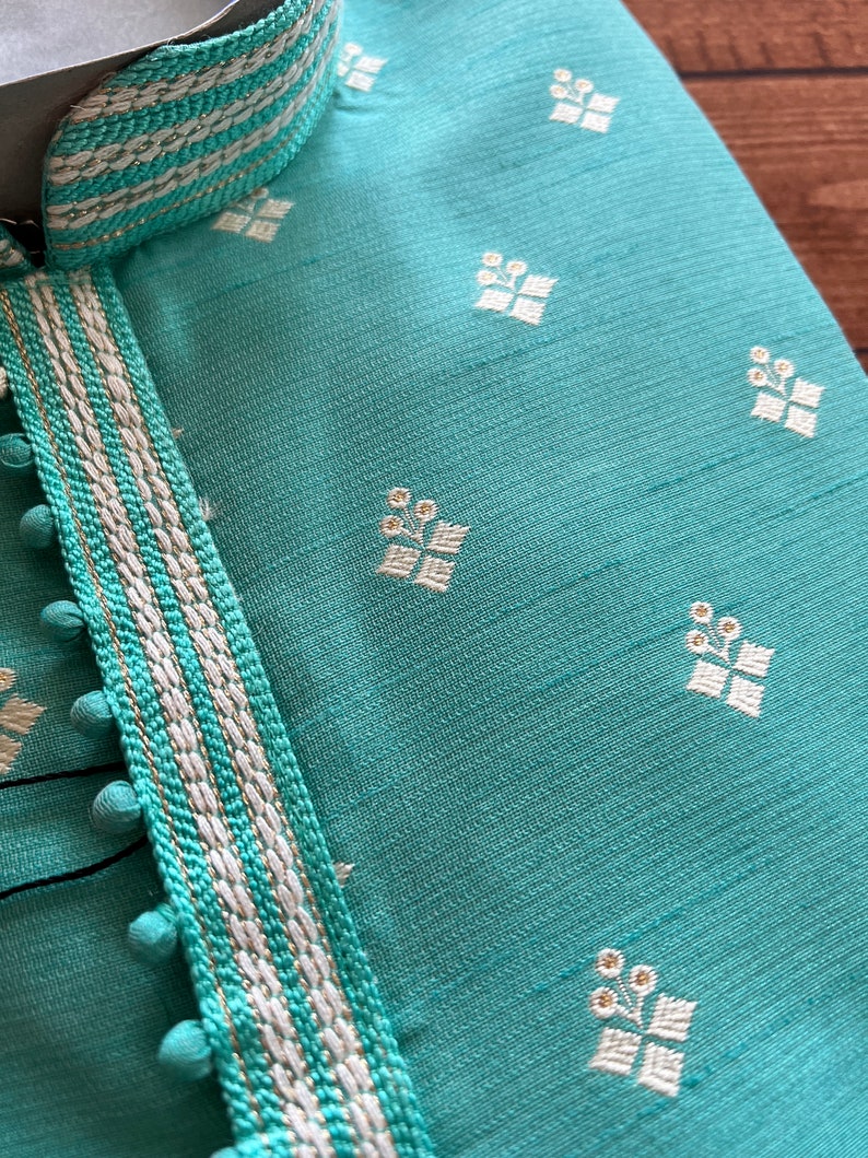Pode incluir: Uma kurta azul turquesa com uma guarni&ccedil;&atilde;o branca e dourada. A guarni&ccedil;&atilde;o tem uma s&eacute;rie de pequenos pompons brancos ao longo da borda. A kurta tem um padr&atilde;o floral branco.