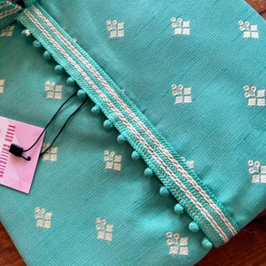 Pode incluir: Uma kurta azul turquesa com um padr&atilde;o bordado branco e dourado. A kurta tem uma bainha branca com detalhes dourados e pompons brancos ao longo das bordas. A etiqueta diz "KASH COLLECTION".